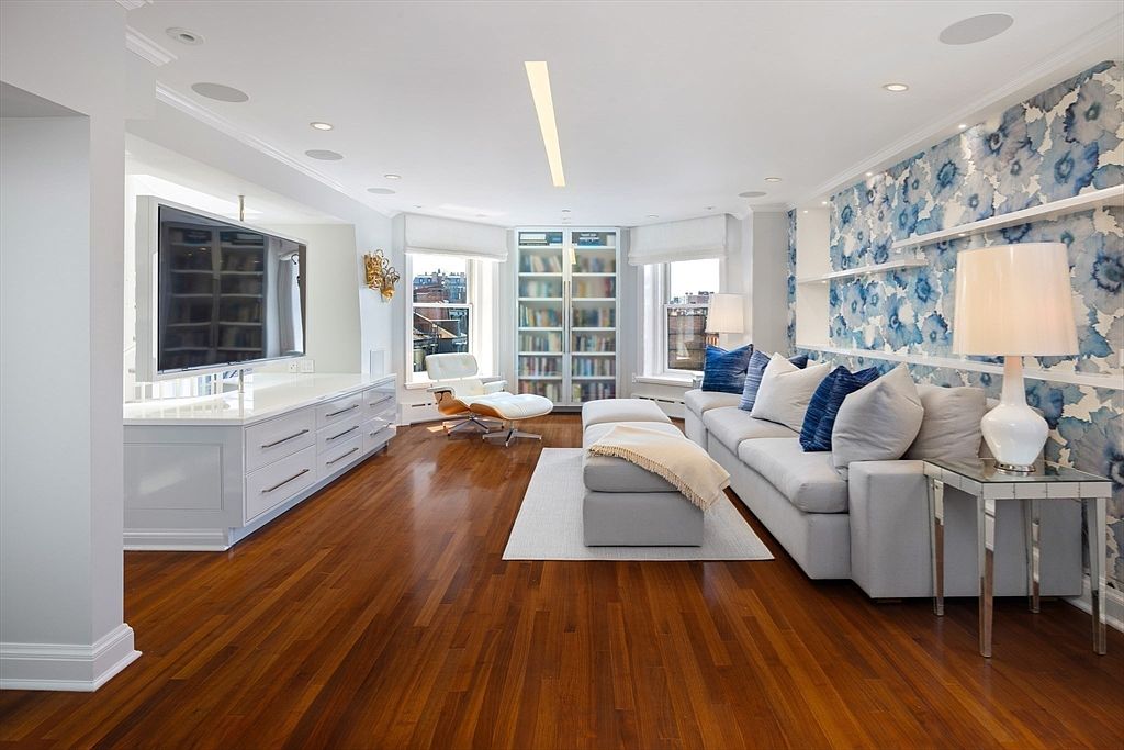 This is a bright and modern living room featuring hardwood floors, a large gray sectional sofa with blue and white pillows, and a custom built-in entertainment center with a large flat-screen TV. The room is well-lit with natural light from the windows and features a unique blue floral wallpaper accent wall. The overall impression is one of sophisticated comfort and style.