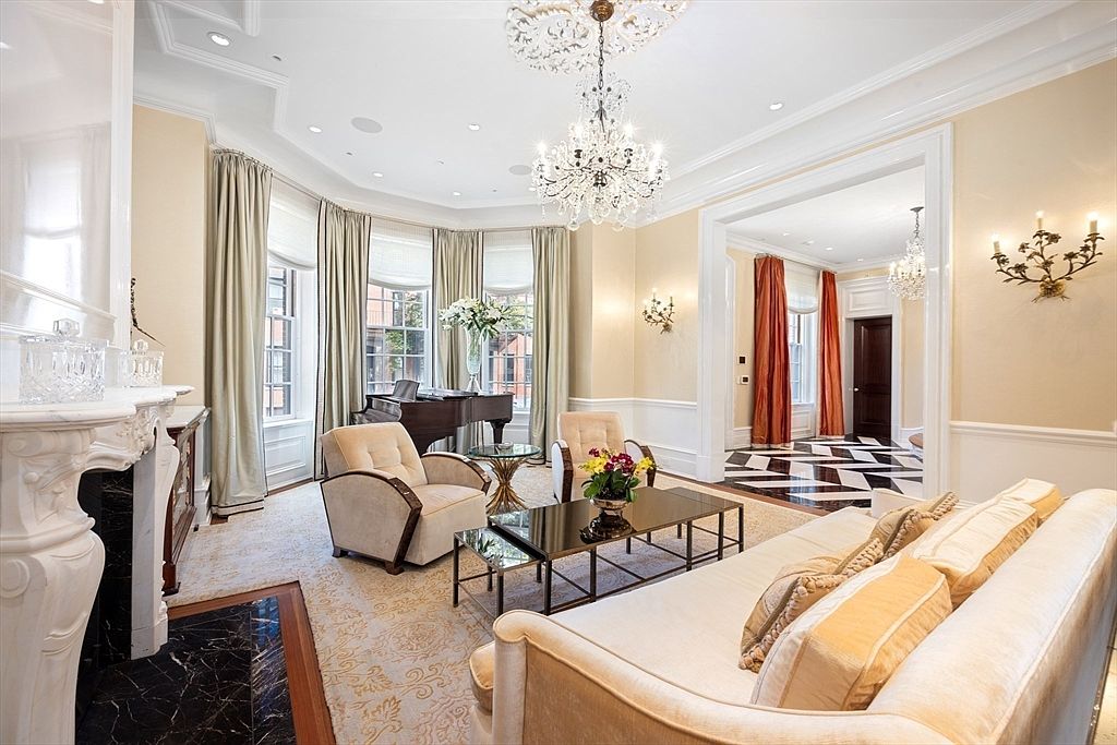 This is an elegant living room featuring a grand piano, a crystal chandelier, and sophisticated furnishings. The room is bathed in soft, natural light from the bay windows, complemented by the warm tones of the walls and the plush seating. The space exudes luxury and comfort, perfect for entertaining or relaxing.