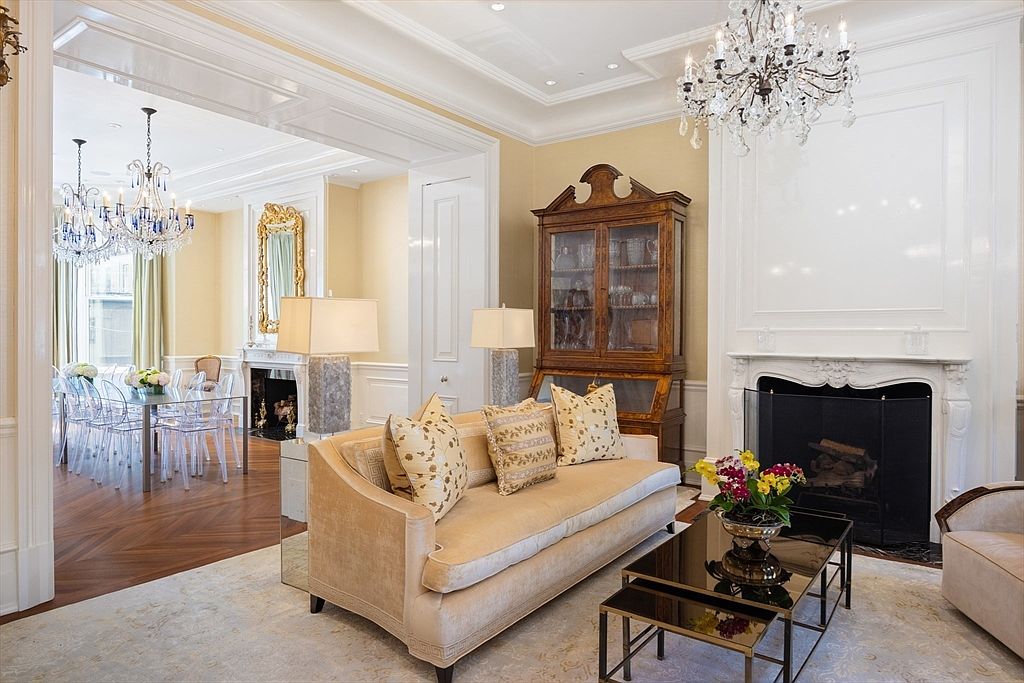 This is an interior shot of an elegant living room featuring a beige sofa with decorative pillows, a dark wood china cabinet, and a white fireplace. A crystal chandelier hangs above, and there is an open doorway leading to a dining area with a glass table and clear chairs. The room exudes a sense of luxury and sophistication.