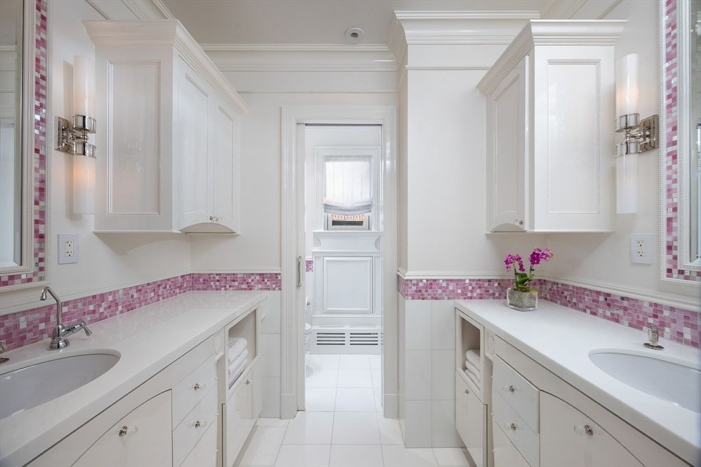 This is a well-lit bathroom featuring dual vanities with white cabinetry and countertops, accented by a pink mosaic tile backsplash. The space is clean and bright, with a doorway leading to another room, suggesting a suite layout. The overall impression is one of elegance and functionality.