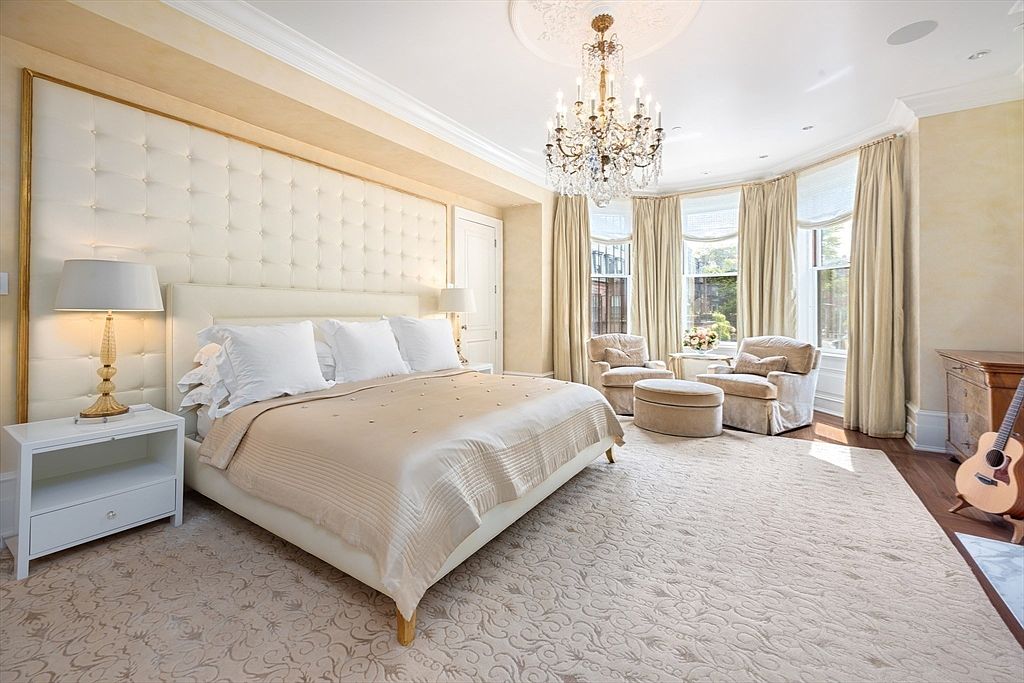 This is a luxurious primary bedroom featuring a large bed with a tufted headboard and a neutral-toned color scheme. The room is well-lit by a chandelier and natural light from the bay windows, which are framed by elegant drapes. The space includes a seating area with armchairs and an ottoman, creating a relaxing and inviting atmosphere.