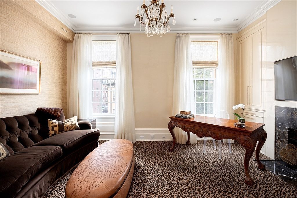 This is an interior shot of a sophisticated home office or study. The room features a brown tufted sofa, a leather ottoman, and a carved wooden desk with clear acrylic chairs. The walls are a mix of textured wallpaper and painted surfaces, complemented by a leopard print carpet, creating a luxurious and stylish workspace.