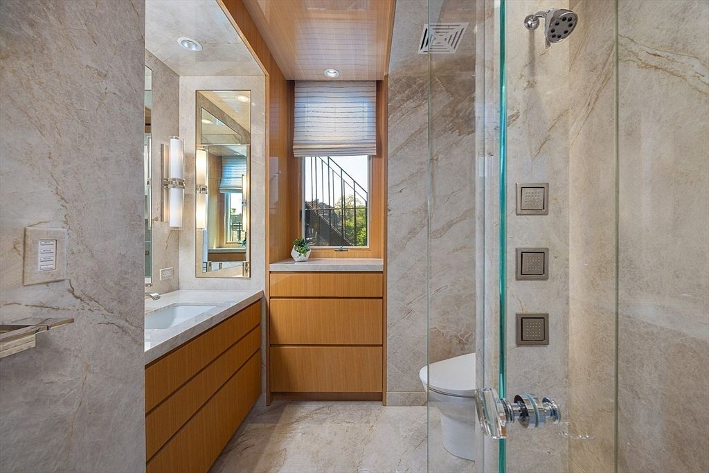 This is a well-lit primary bathroom featuring marble walls and flooring, creating a luxurious and elegant atmosphere. The bathroom includes a vanity with wooden drawers, a large mirror, and a modern shower with glass doors and multiple shower heads. A window with a blind provides natural light and a view to the outside.