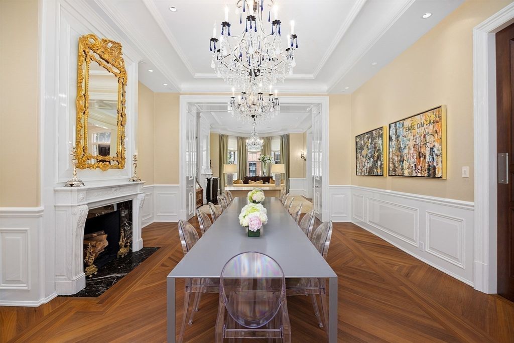 This is an elegant dining room featuring a long gray table with clear chairs, positioned under a multi-tiered crystal chandelier. The room is decorated with classic wainscoting, light yellow walls, and hardwood floors laid in a herringbone pattern. A decorative fireplace with a gold-framed mirror above it adds a touch of traditional charm, while artwork on the opposite wall provides a modern contrast.