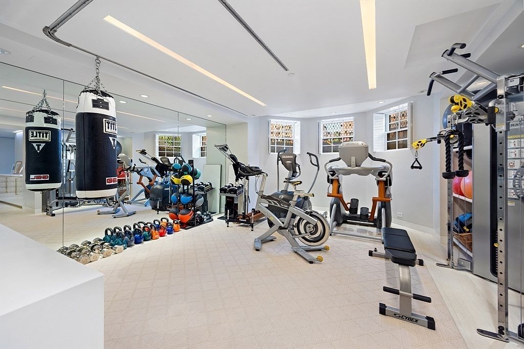 This is a well-equipped home gym featuring a variety of exercise equipment including punching bags, free weights, stationary bikes, and a multi-station weight machine. The room is bright and spacious, with mirrored walls and recessed lighting, creating an inviting and motivating workout environment. The neutral color palette and clean design contribute to a modern and functional space.