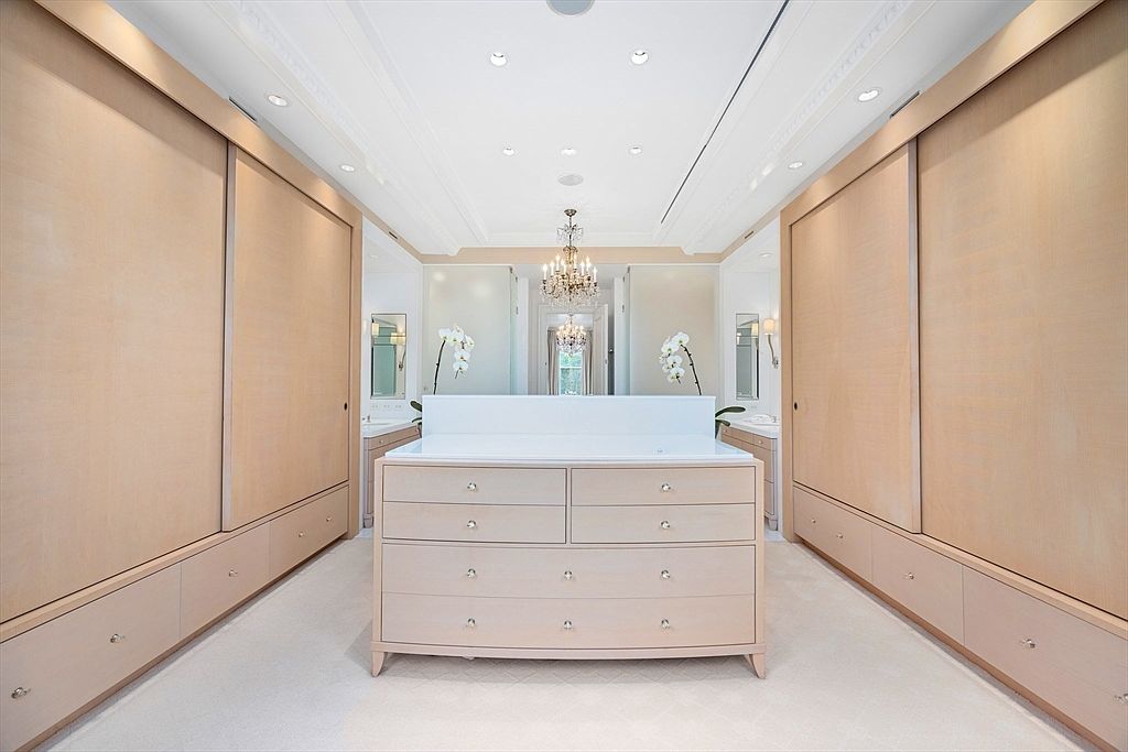 This is a well-organized walk-in closet featuring custom cabinetry with light wood finishes and sleek hardware. The closet includes ample storage with drawers and sliding doors, and a central dresser provides additional surface space. A chandelier adds a touch of elegance to the space, which is brightly lit and appears spacious.