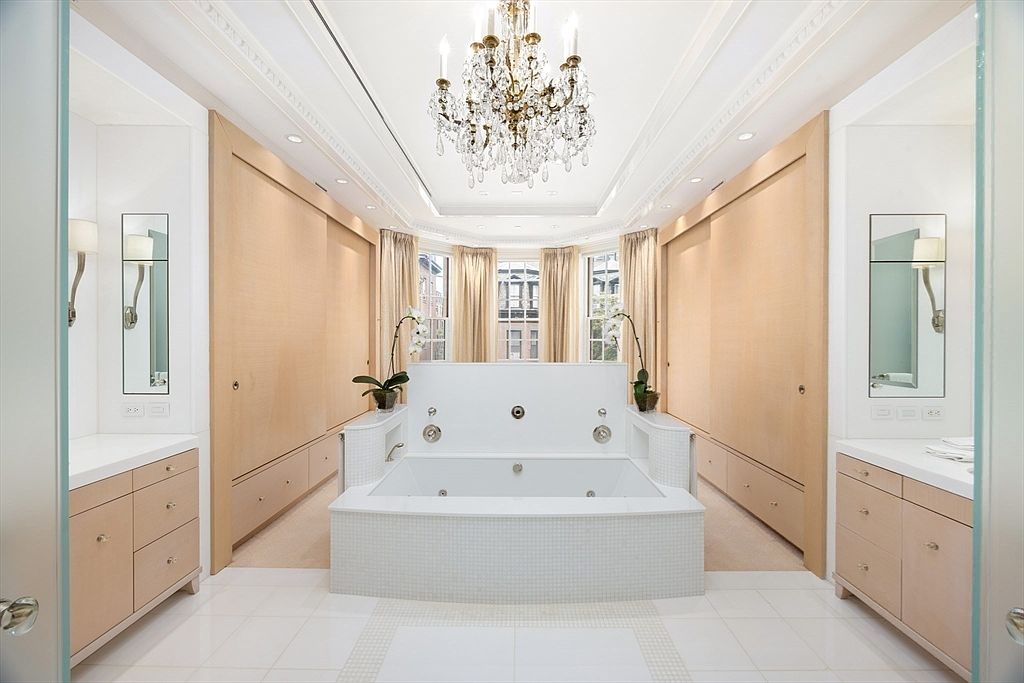 This is a luxurious primary bathroom featuring a large, tiled jacuzzi tub as the focal point, flanked by matching vanities with light wood cabinetry and mirrors. The room is illuminated by a crystal chandelier and recessed lighting, with natural light filtering through the windows behind the tub. The overall impression is one of elegance and relaxation.