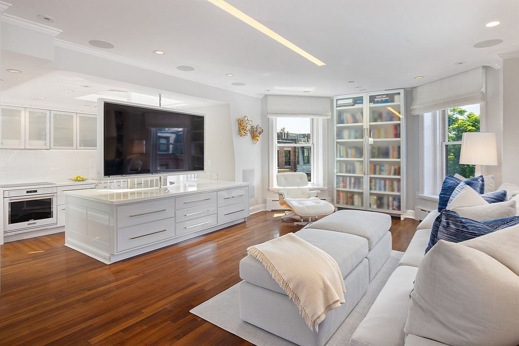 This is a bright and airy living room featuring hardwood floors, a large sectional sofa with throw pillows and a blanket, and a modern bookcase. A unique TV setup is integrated into a kitchen island, and natural light streams in through the windows. The overall impression is one of contemporary elegance and comfort.