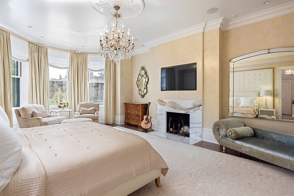 This is a luxurious primary bedroom featuring a large bed with a neutral-toned comforter, a sitting area by a bay window with elegant curtains, and a fireplace with a mounted television above. A large mirror reflects the room, enhancing the sense of space, while a crystal chandelier adds a touch of opulence. The room is decorated in soft, warm tones, creating a serene and inviting atmosphere.