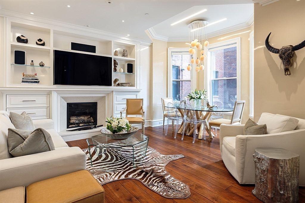 This is an interior shot of a well-decorated living room featuring a fireplace with a mounted television above it, built-in shelving, and a dining area with a glass table. The room is furnished with a white sofa, an armchair, and a zebra-print rug, creating a blend of modern and eclectic styles. The hardwood floors and natural light enhance the inviting atmosphere of the space.