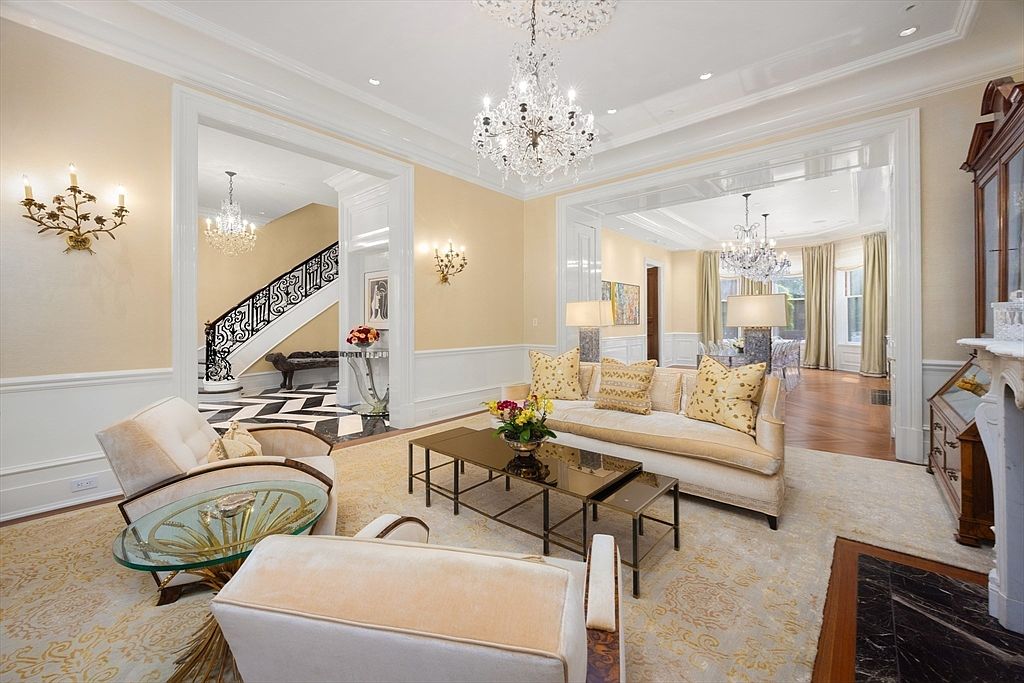 This elegant living room features a sophisticated design with a crystal chandelier, classic furnishings, and detailed architectural moldings. The room opens to a hallway with a staircase and another room with a dining area, creating a sense of spaciousness and flow. The color palette is soft and inviting, enhancing the luxurious feel of the space.