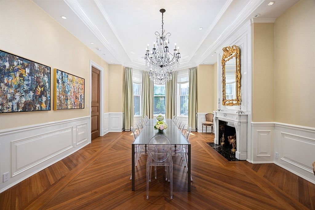 This is an elegant dining room featuring a long, modern dining table with clear chairs, positioned under a sparkling chandelier. The room is decorated with wainscoting, artwork, and a decorative fireplace with a gilded mirror above. The hardwood floors are laid in a herringbone pattern, adding to the room's sophisticated ambiance.