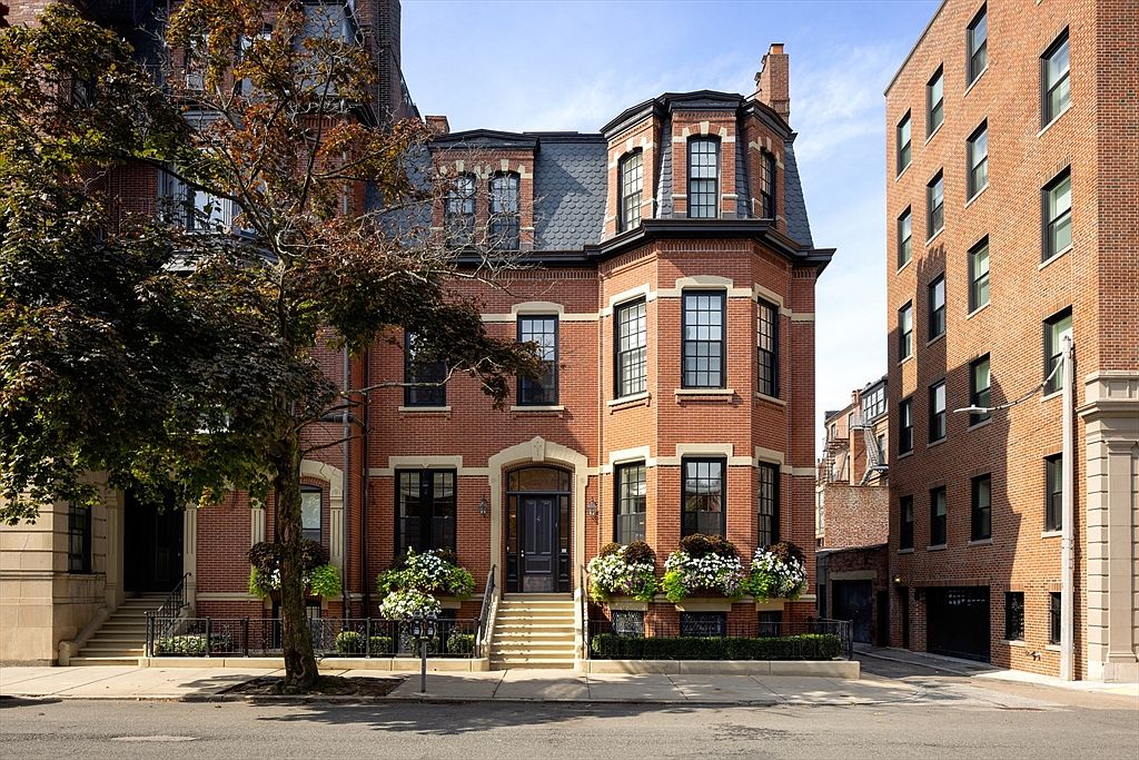 This is a front view of a stately brick townhouse with a prominent bay window section. The building features elegant architectural details, including stone accents around the windows and entryway, and well-maintained landscaping with flower boxes. The overall impression is one of classic elegance and urban sophistication.