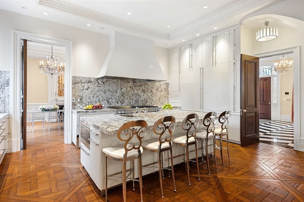 This is a bright and elegant kitchen featuring a large island with seating, white cabinetry, and a decorative backsplash. The hardwood floors are laid in a herringbone pattern, adding a touch of classic charm. The kitchen opens to other rooms, showcasing the home's spacious layout and luxurious design.