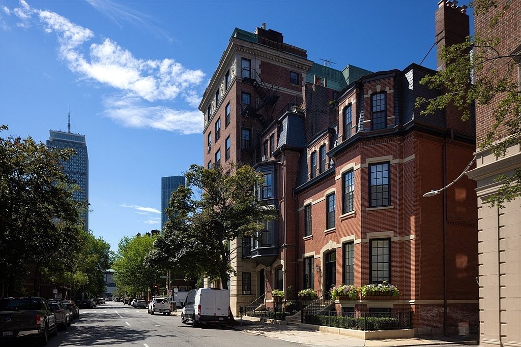 This image showcases the front exterior of a classic brick townhouse in an urban setting. The architecture features detailed brickwork, black-framed windows, and a mansard roof, exuding a sense of timeless elegance. The street view includes mature trees and glimpses of modern skyscrapers in the distance, blending historic charm with city life.