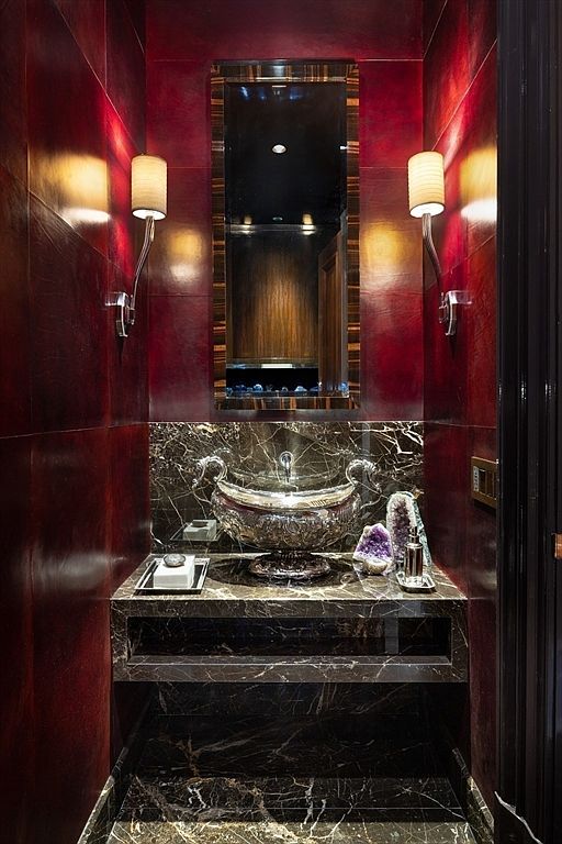 This is a luxurious guest bathroom featuring rich, dark red walls and a striking black marble vanity. The sink is an ornate, silver vessel, complemented by elegant sconces and a decorative mirror. The space exudes sophistication and opulence, making it a memorable feature of the home.
