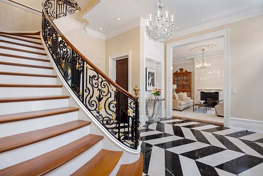 This is an interior shot of a grand foyer featuring a curved staircase with ornate wrought iron railings and wood treads. The black and white geometric tile flooring adds a modern touch, while a crystal chandelier provides elegant lighting. The space opens into a living area with a fireplace, creating a luxurious and inviting atmosphere.