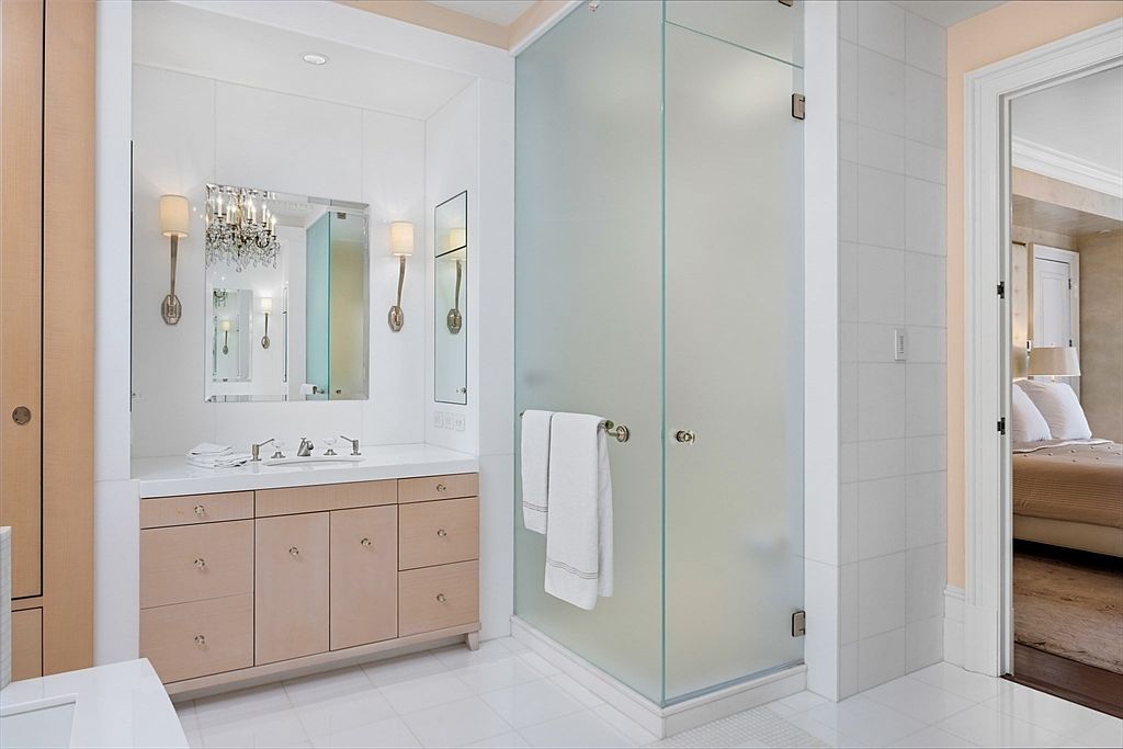 This is a well-lit primary bathroom featuring a vanity with light wood cabinetry, a white countertop, and a large mirror with sconces. A glass-enclosed shower is adjacent to the vanity, and a doorway leads to the bedroom. The bathroom has a clean and elegant aesthetic with white tile flooring and walls.