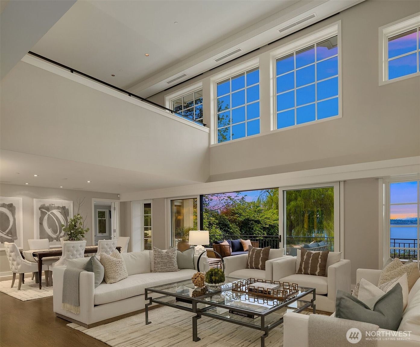 This is a spacious and luxurious living room featuring high ceilings and large windows that offer ample natural light and views of the outdoors. The room is furnished with comfortable white sofas, a glass coffee table, and a neutral-toned rug, creating an inviting and elegant atmosphere. The open floor plan seamlessly connects the living area to the dining space, enhancing the sense of spaciousness and flow.