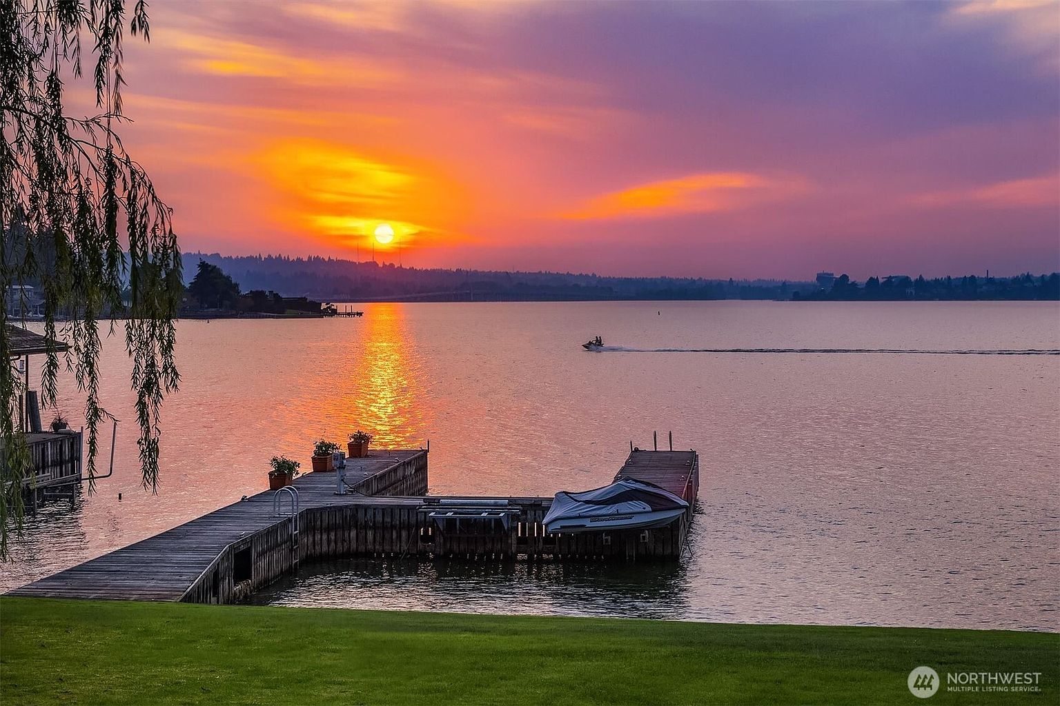 The image showcases a serene waterfront property at sunset. A wooden dock extends into the calm water, featuring potted plants and a boat lift with a boat. The vibrant sunset paints the sky with hues of orange and purple, creating a picturesque and inviting scene.