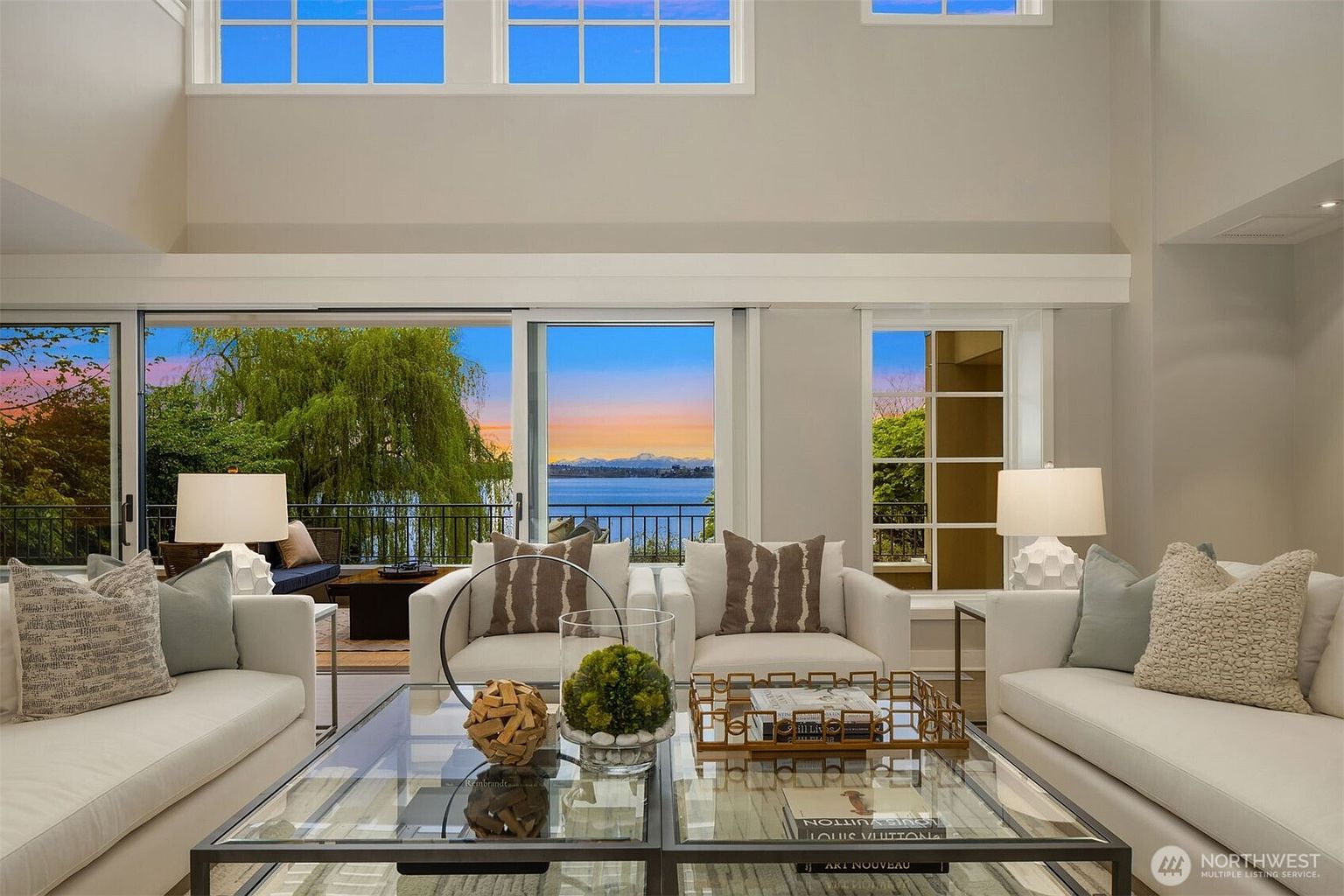 This is a bright and airy living room with a high ceiling and large windows offering a scenic view of the water and sunset. The room is furnished with white sofas and chairs, creating a clean and modern aesthetic. A glass coffee table sits in the center, adding to the room's sophisticated ambiance.