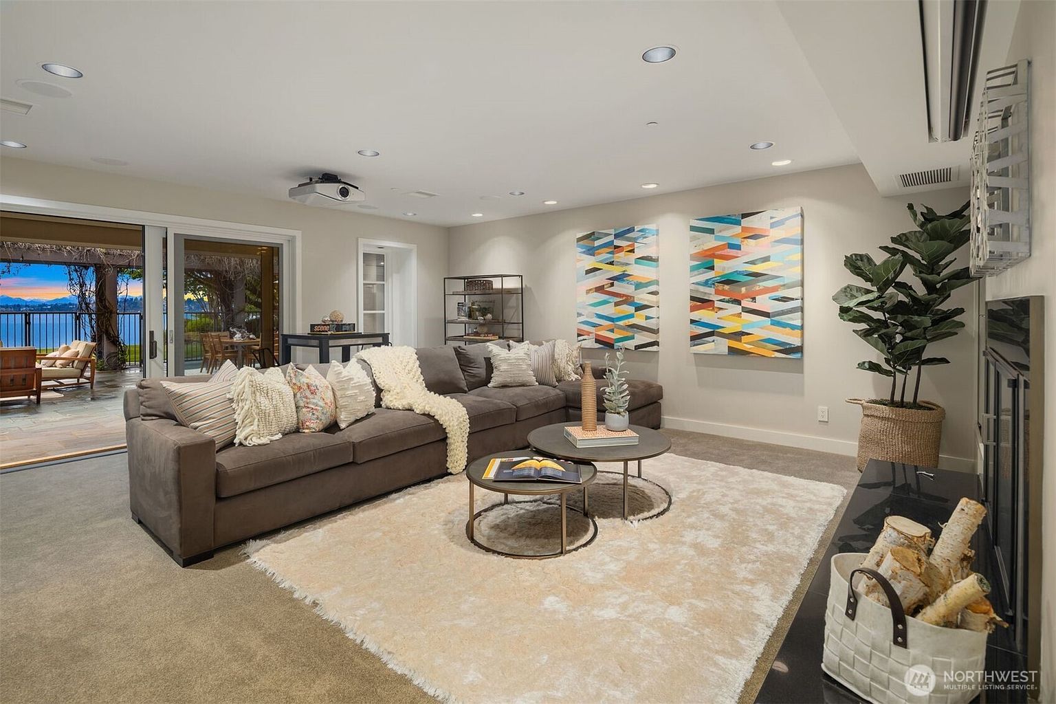 This is a cozy and inviting living room featuring a large sectional sofa with decorative pillows, two round coffee tables, and a plush area rug. The room is well-lit with recessed lighting and natural light from the sliding glass doors that lead to an outdoor patio with a view of the water. Artwork adorns the walls, and a fireplace with a basket of logs adds to the room's warmth and character.