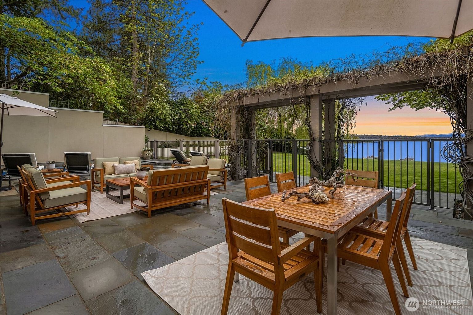 This image showcases a luxurious outdoor patio area, complete with wooden furniture arranged for lounging and dining. The patio features a stone floor and is partially covered by a pergola draped with vines, offering a view of a serene body of water at sunset. The space is designed for relaxation and entertainment, highlighting the property's outdoor living potential.