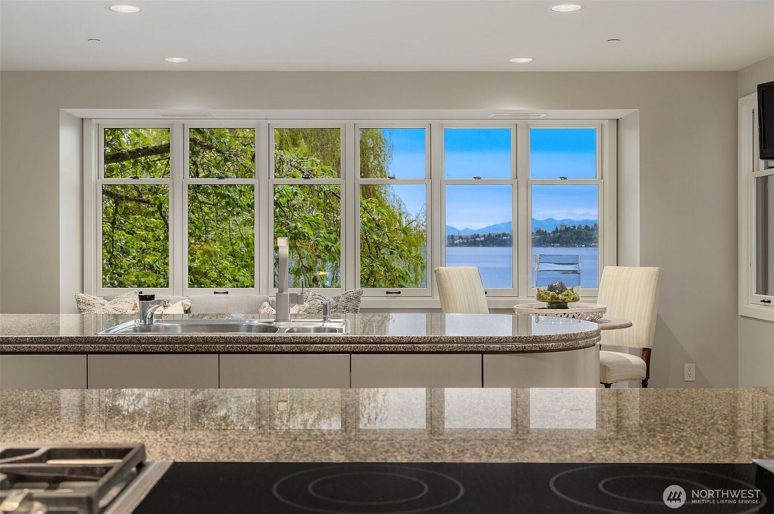 This is a bright kitchen featuring a large window with a view of the water and mountains. The kitchen has a granite countertop and a stainless steel sink. The overall impression is clean and modern, with a focus on the view.