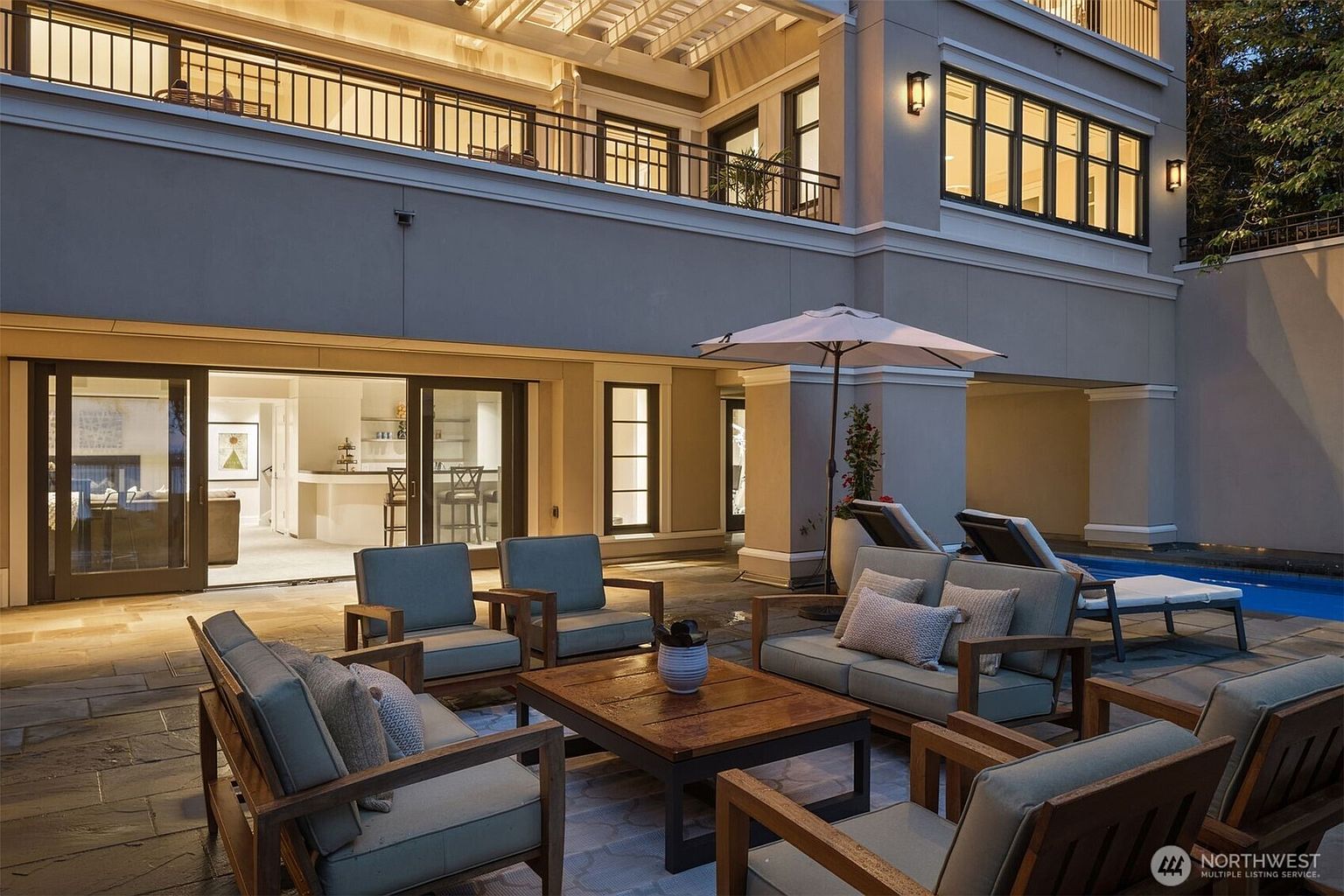 This image showcases a luxurious outdoor patio area, featuring comfortable lounge seating arranged around a wooden coffee table. The patio is paved with stone and includes an umbrella for shade, with a glimpse of a swimming pool in the background. The space is adjacent to the building, with large sliding doors leading to the interior, creating a seamless indoor-outdoor living experience.