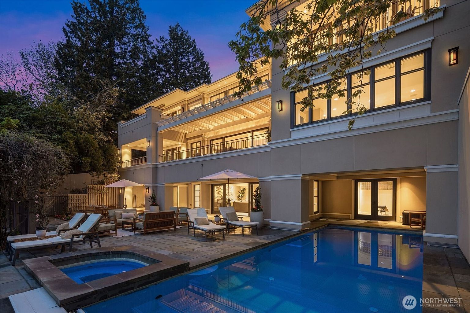 This image showcases the rear exterior of a luxurious home, highlighting a stunning pool and spa area. The multi-level patio features lounge chairs, umbrellas, and outdoor seating, creating an inviting space for relaxation and entertainment. The architecture of the home is modern with clean lines, large windows, and a covered balcony, all illuminated by warm lighting, enhancing the property's appeal.