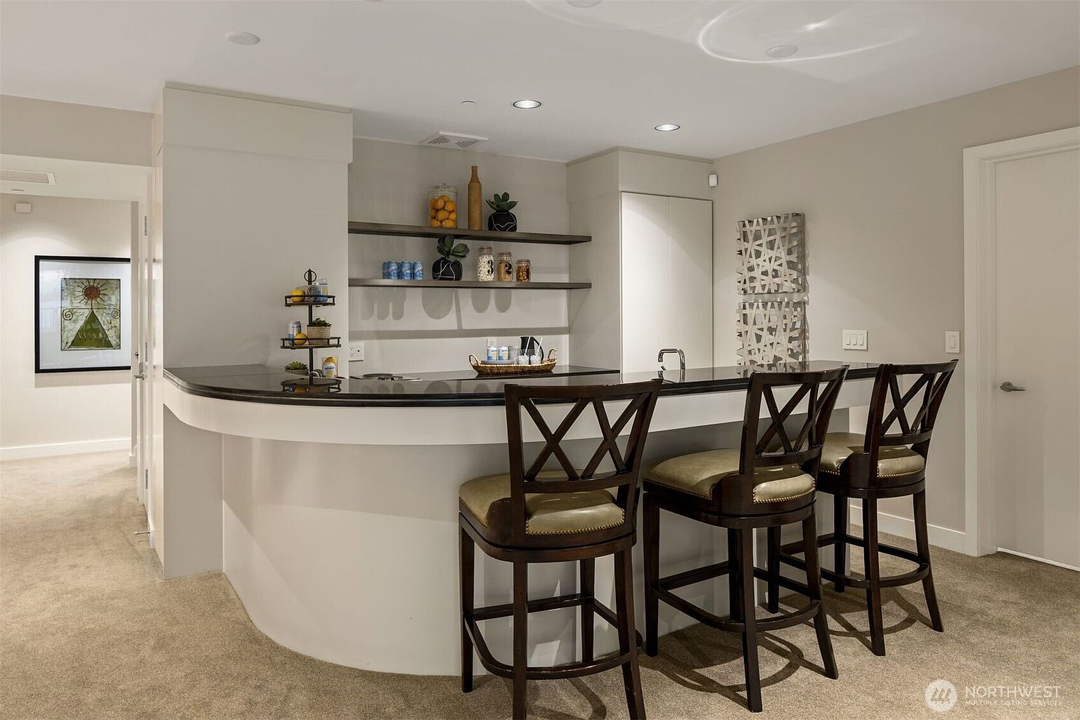 This interior shot showcases a stylish basement bar area. The bar features a curved white base with a dark countertop, complemented by three dark wood bar stools with light-colored cushions. Open shelving displays decorative items, and the overall ambiance is modern and inviting, perfect for entertaining.