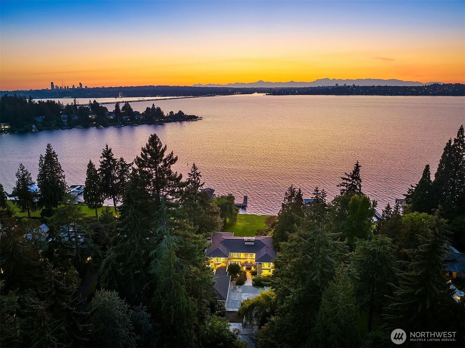This aerial view showcases a luxurious waterfront estate at sunset. The large home is nestled among mature trees, with a sprawling lawn leading to the water's edge. The warm glow of the setting sun reflects on the lake, creating a serene and picturesque setting, while the city skyline is visible in the distance.