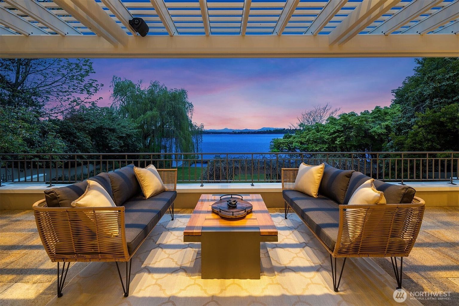 This image showcases a luxurious outdoor patio or deck area, featuring comfortable seating arrangements with stylish sofas and throw pillows. A modern coffee table sits in the center, and the space is framed by a scenic view of a lake or ocean at dusk. The overall impression is one of relaxation and upscale outdoor living.