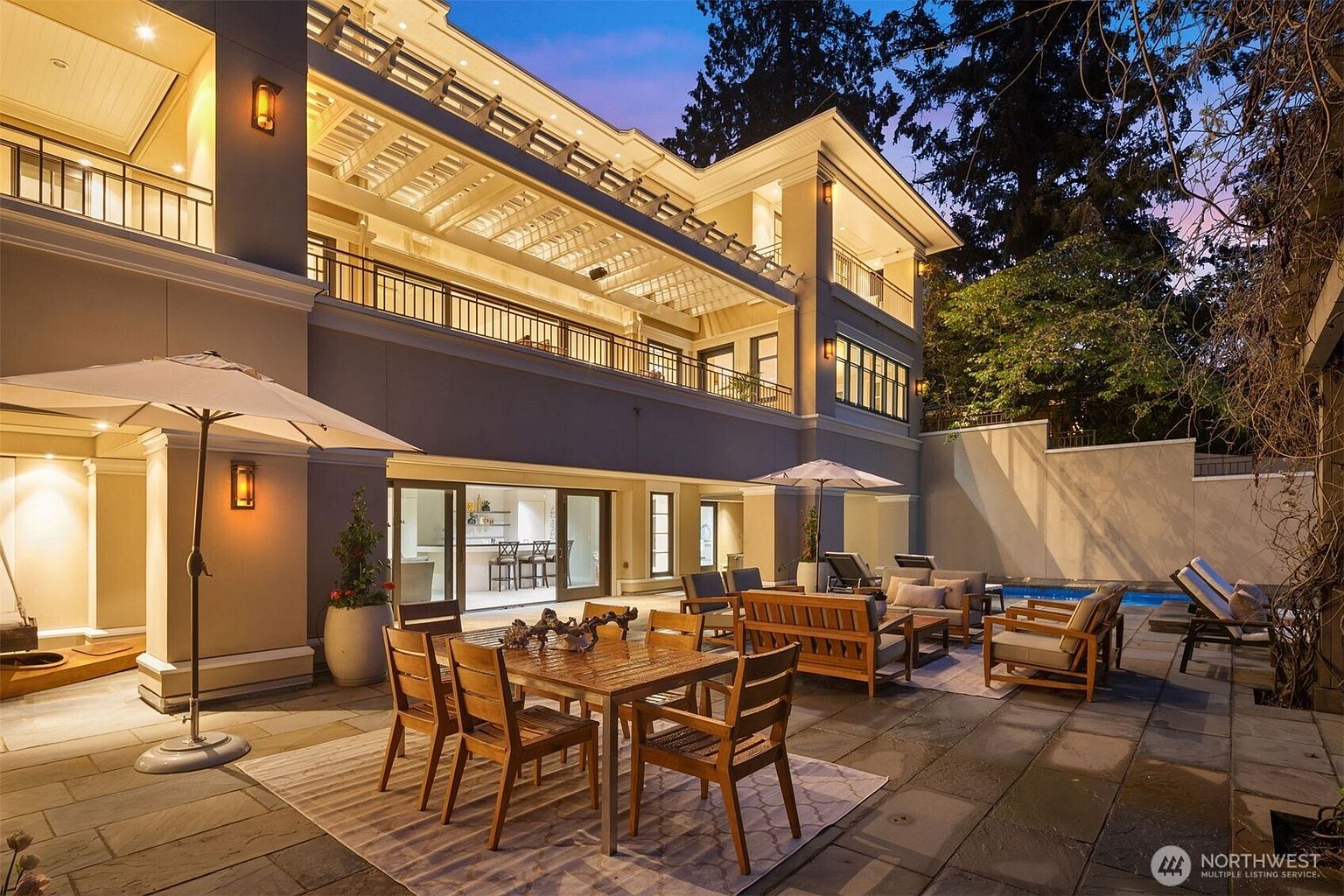 This image showcases a luxurious outdoor patio area, featuring elegant wooden furniture arranged for dining and lounging. The patio is paved with stone tiles and includes a large umbrella for shade. The backdrop is a well-maintained two-story building with a balcony and a glimpse of a swimming pool, creating an inviting and sophisticated outdoor living space.