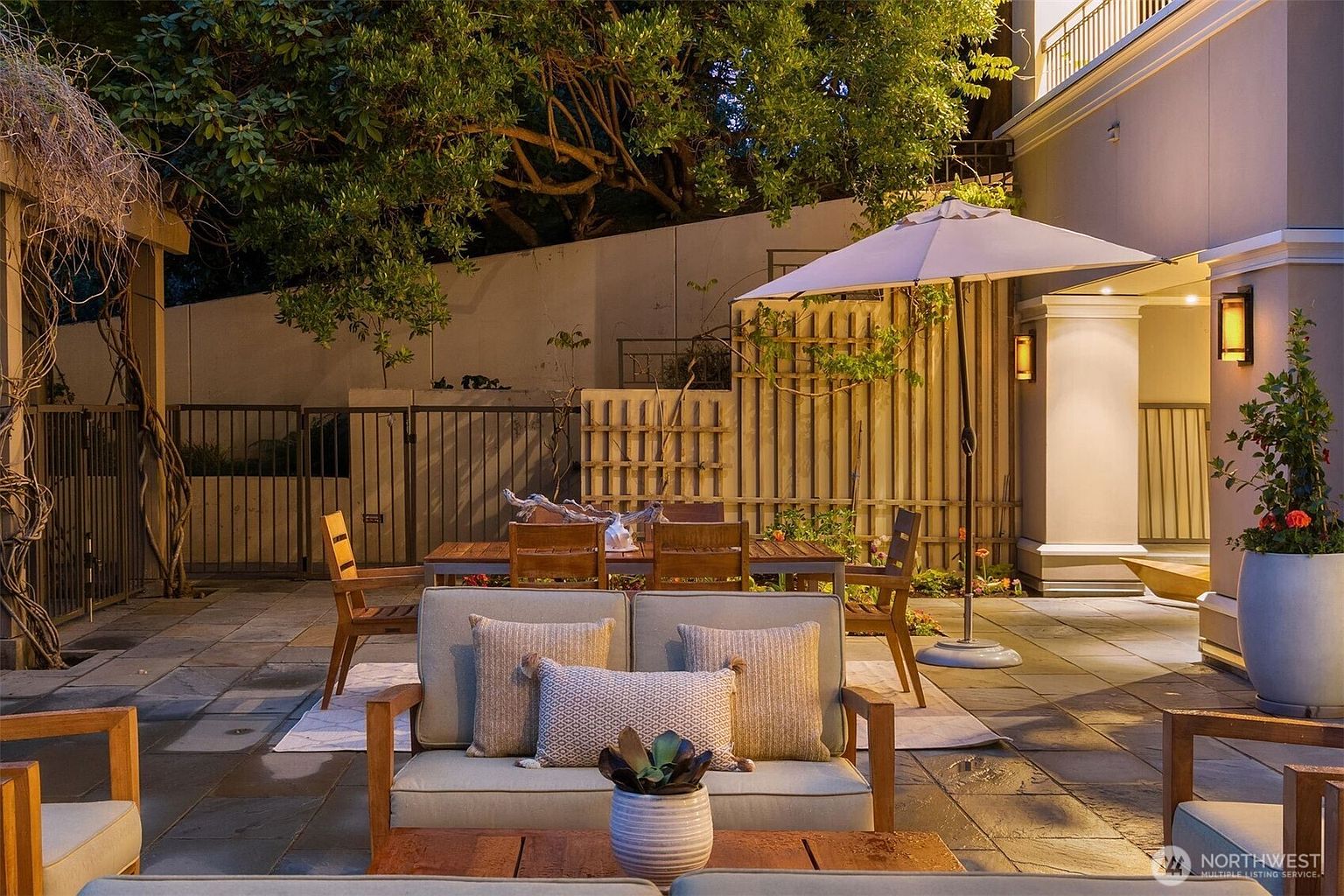 This image showcases a beautifully designed outdoor patio area, perfect for relaxation and entertaining. The space features comfortable seating arrangements, a dining table set under a large umbrella, and tasteful landscaping. The lighting creates a warm and inviting ambiance, highlighting the quality of the outdoor furniture and the overall design.