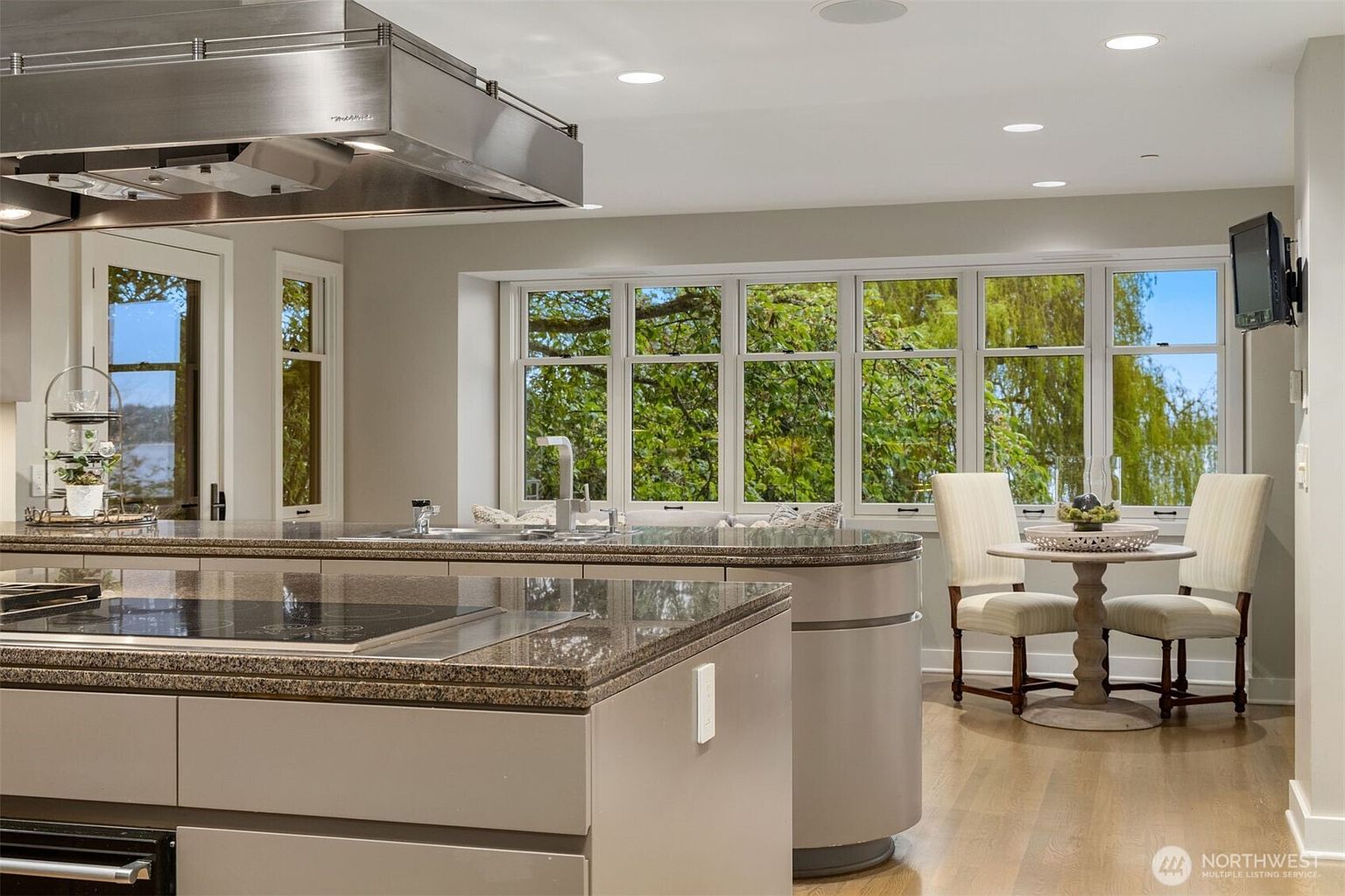 This is a well-lit kitchen featuring a large island with a cooktop and granite countertops. A stainless steel range hood hangs above the island. A breakfast nook with a round table and two chairs sits near a large window offering a view of lush greenery.