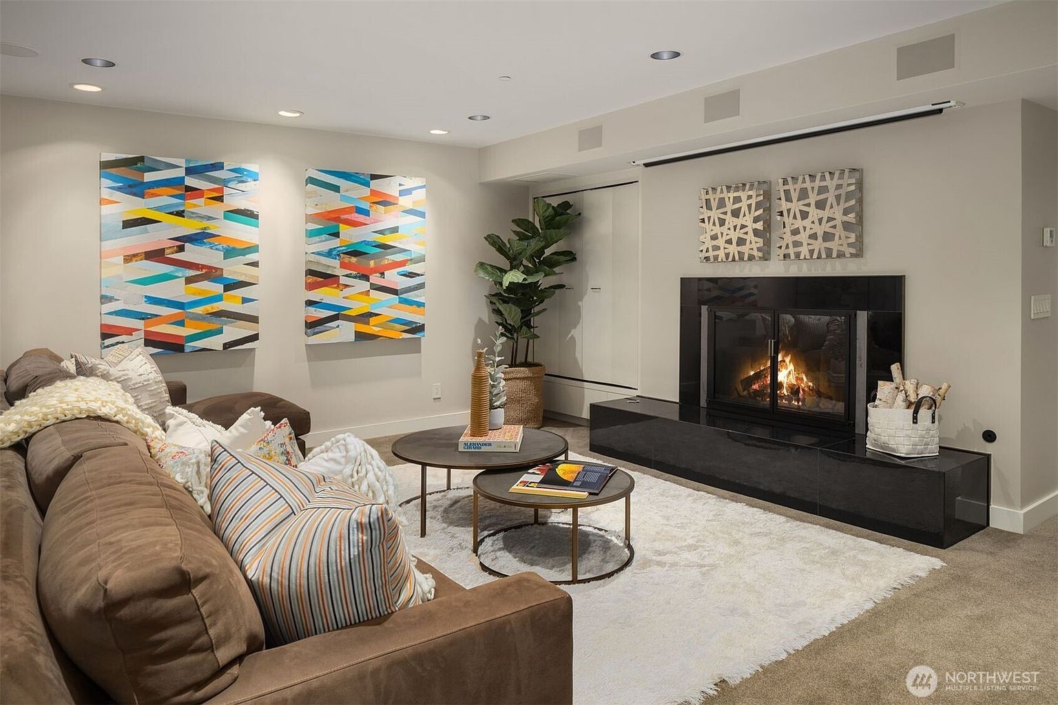 This is an inviting living room featuring a comfortable brown sectional sofa adorned with decorative pillows and a throw blanket. The room is decorated with modern art pieces and a fireplace, creating a cozy and stylish atmosphere. The neutral color palette and soft lighting enhance the room's warmth and appeal.