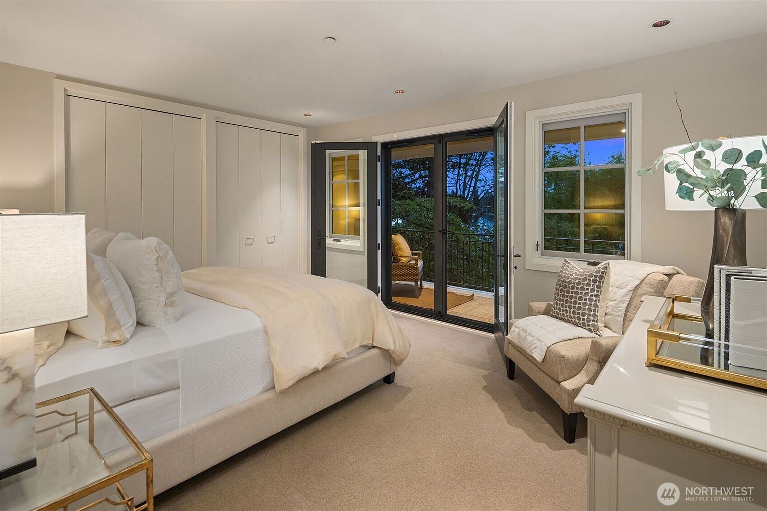 This is a well-lit primary bedroom featuring a neutral color palette. The room includes a bed with soft linens, built-in wardrobes, and a seating area by a window. French doors lead to a balcony, offering an indoor-outdoor living experience.