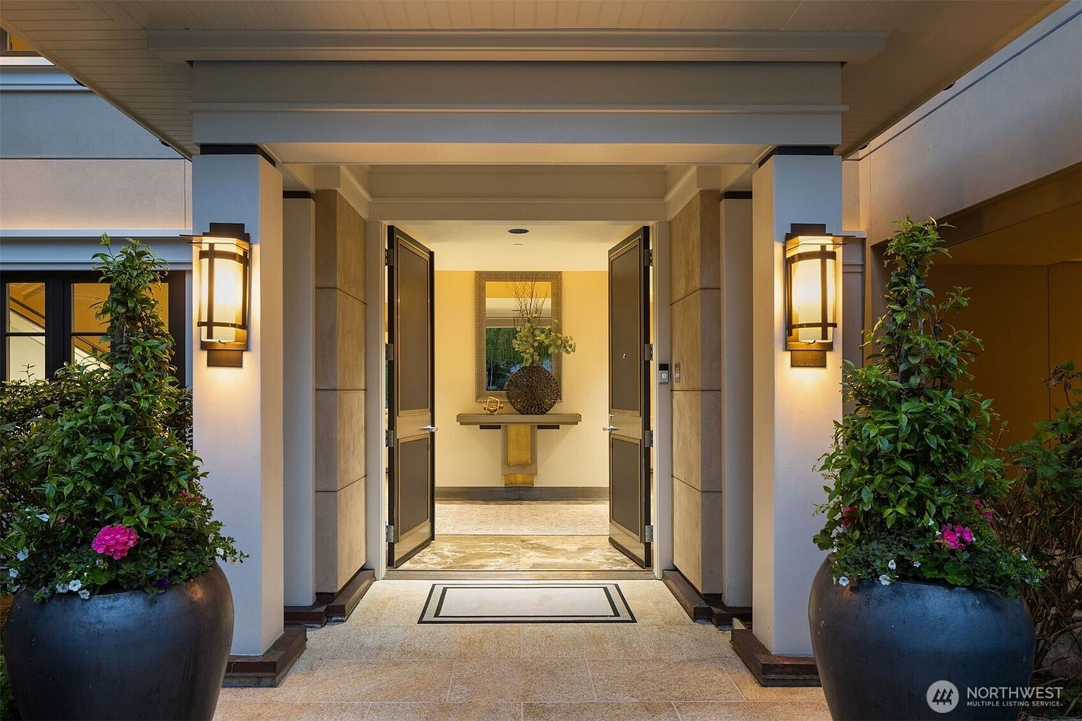 The image showcases a grand entryway to a home, featuring double doors flanked by stone pillars with sconce lighting. Large potted plants add a touch of greenery and elegance to the entrance. The flooring is made of light-colored stone tiles, creating a welcoming and luxurious first impression.