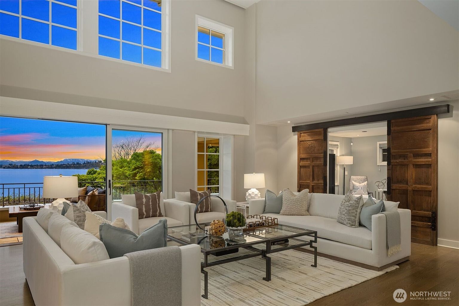 This is a spacious living room with high ceilings and large windows offering a scenic view. The room features white sofas, a glass coffee table, and a light-colored rug. A unique architectural element is the large wooden sliding barn doors, adding a rustic touch to the modern interior.