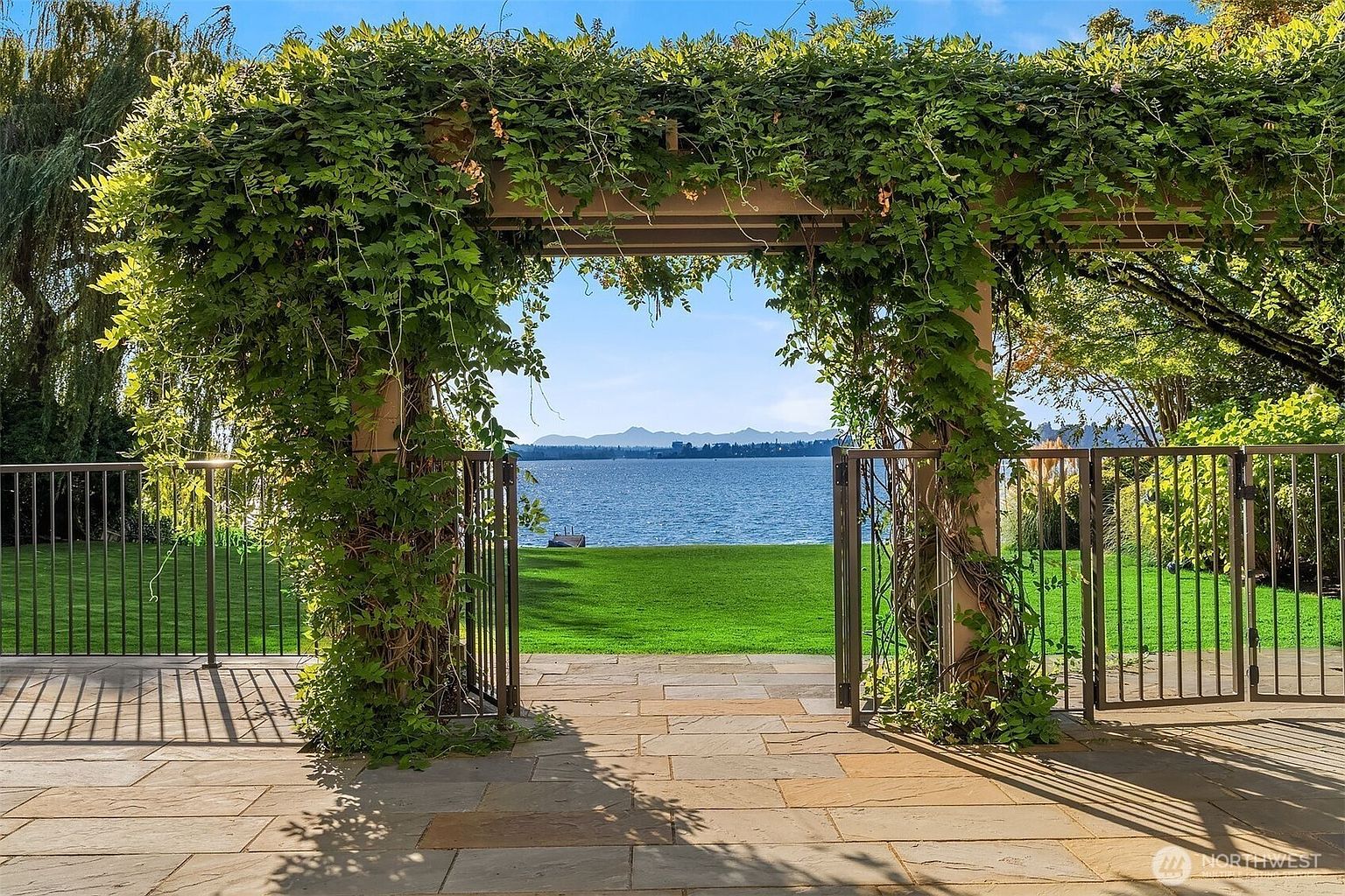 This image showcases a beautifully landscaped yard with a stone patio leading to a lush green lawn and a serene lake view. An arched pergola covered in climbing vines frames the scene, creating an inviting and picturesque outdoor space. A metal fence encloses the yard, adding a touch of elegance and security.