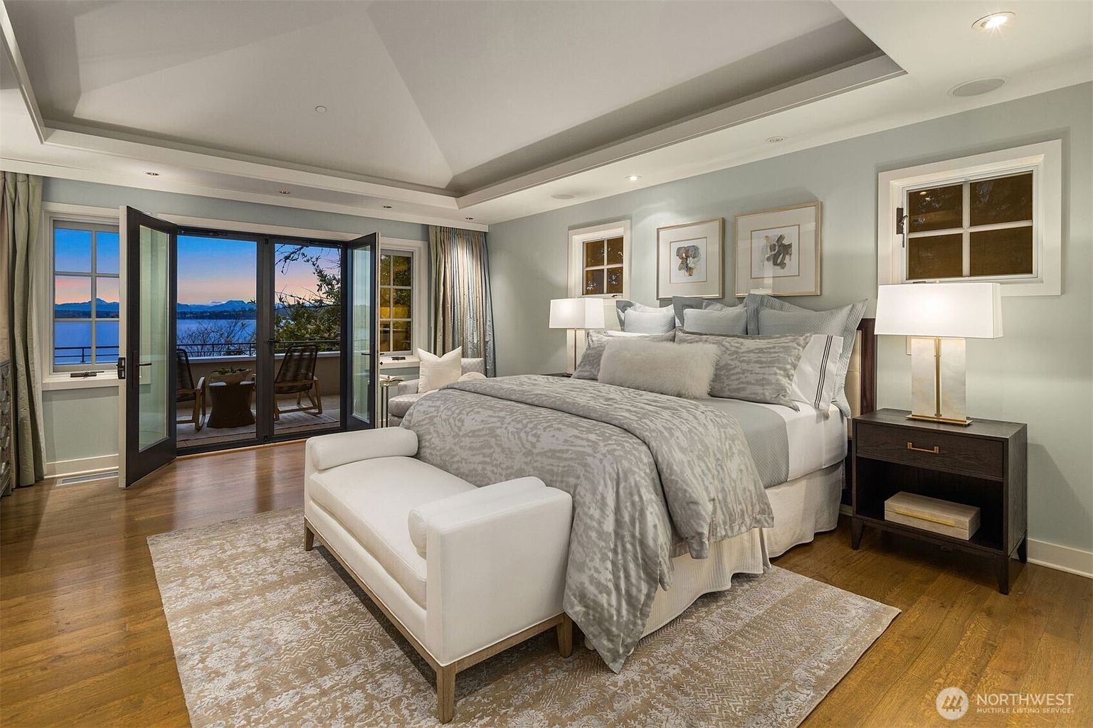 This is a luxurious primary bedroom featuring a king-size bed with a patterned gray comforter and multiple pillows. The room has a tray ceiling, light blue walls, and large windows and doors that open to a balcony with a water view. A white bench sits at the foot of the bed, and a dark wood nightstand with a lamp is on one side.