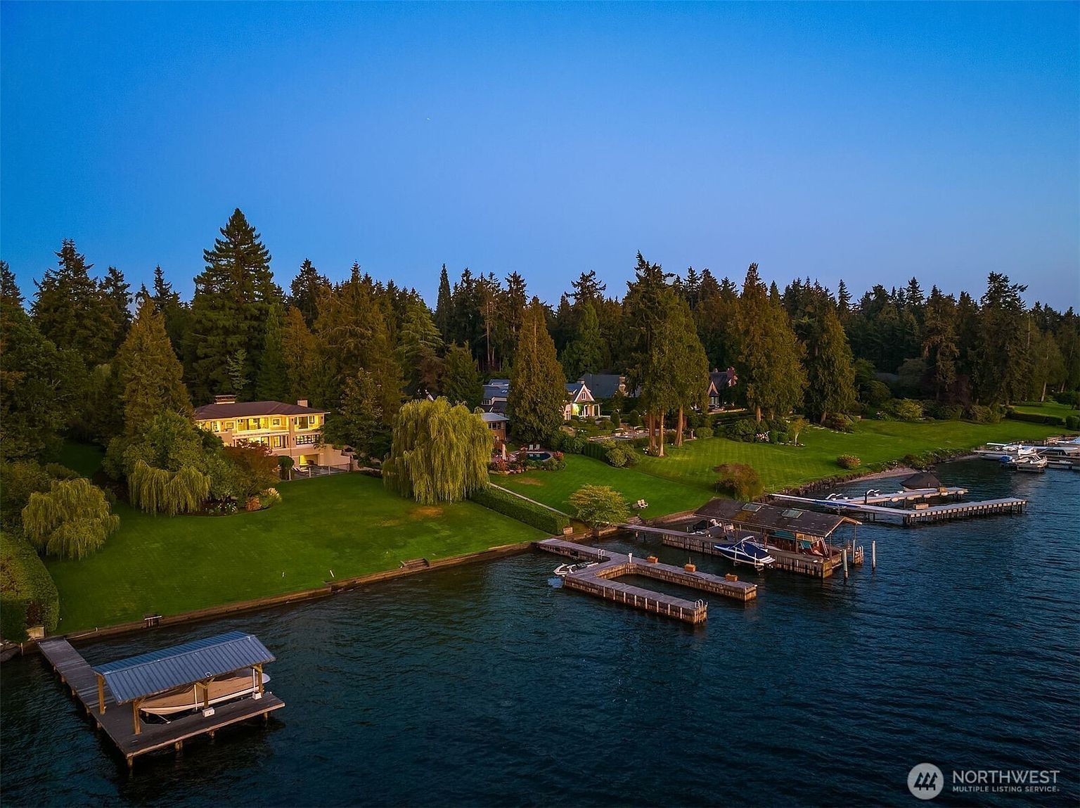 This aerial shot showcases a stunning waterfront property featuring lush green lawns, mature trees, and multiple docks extending into the calm, dark blue water. Several houses are visible, suggesting a luxurious lakeside community. The image captures a serene and upscale living environment, perfect for boating enthusiasts.