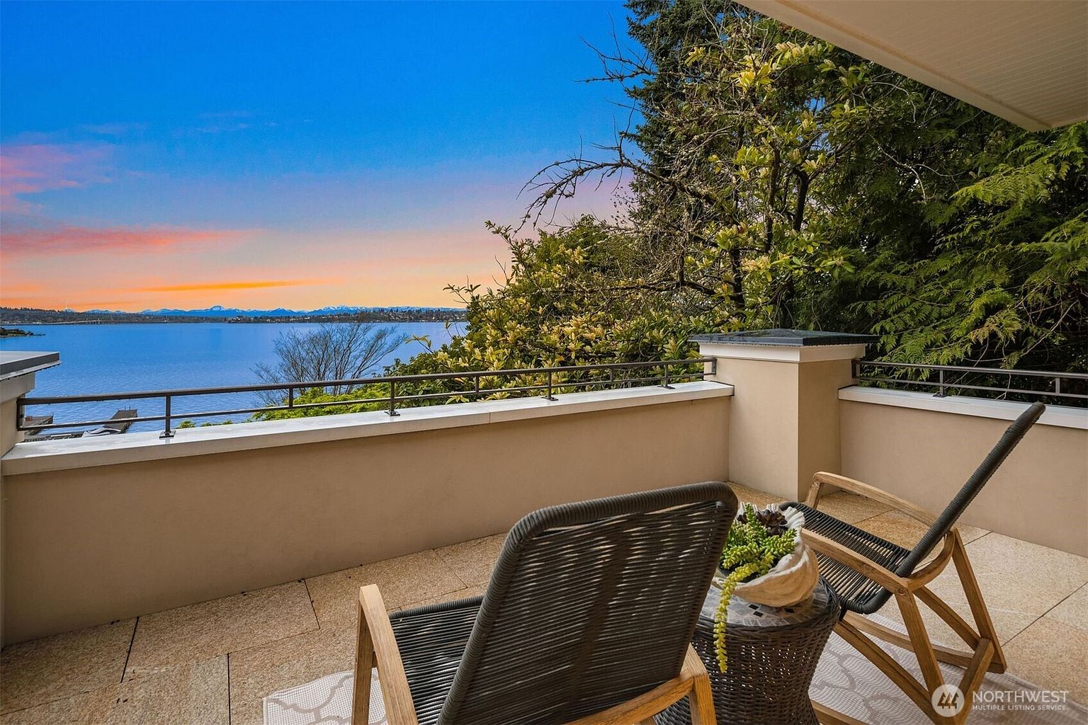This image showcases a serene balcony setting with two modern chairs and a small table, offering a stunning view of the water and distant mountains at sunset. The balcony features a neutral-toned wall and tiled flooring, creating a relaxing outdoor living space. Lush greenery surrounds the balcony, enhancing the sense of privacy and connection with nature.