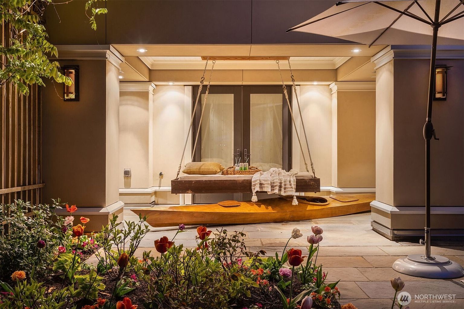 This image showcases a beautifully designed outdoor patio area. A unique swing bed hangs under a covered section, complemented by a wooden kayak resting beneath it. The foreground features a vibrant flower garden, adding a touch of nature to the sophisticated outdoor living space.