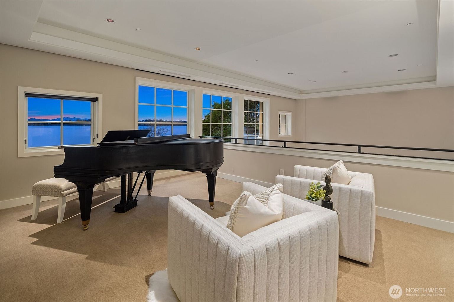 This is an interior shot of a living room featuring a grand piano near a window with a scenic view. Two modern, white armchairs are positioned in the foreground, creating a cozy seating area. The room has a neutral color palette, with beige walls and carpet, contributing to a serene and elegant atmosphere.
