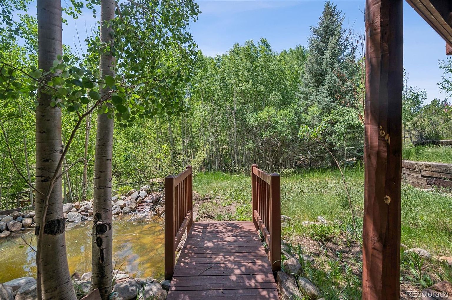 This image showcases a serene yard or garden setting, featuring a small wooden bridge leading towards a lush green area with various trees and foliage. A small pond is visible to the left, adding to the natural ambiance. The scene evokes a sense of tranquility and privacy, highlighting the property's outdoor appeal.