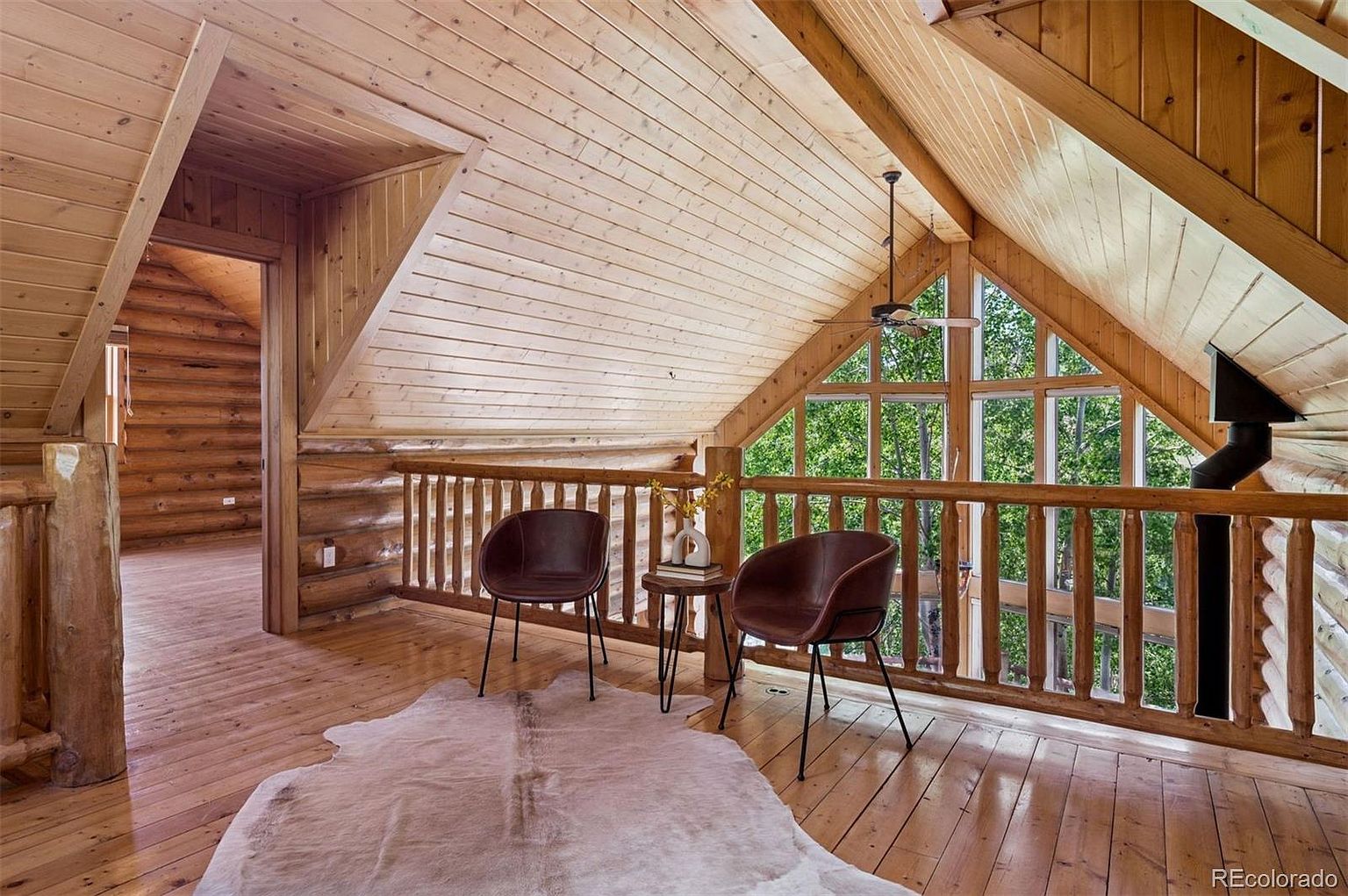 This image showcases a cozy attic space featuring natural wood finishes on the walls, ceiling, and railing. Two chairs and a small table create a seating area near a large window offering a view of the surrounding trees. The space is well-lit and inviting, with a rustic charm.