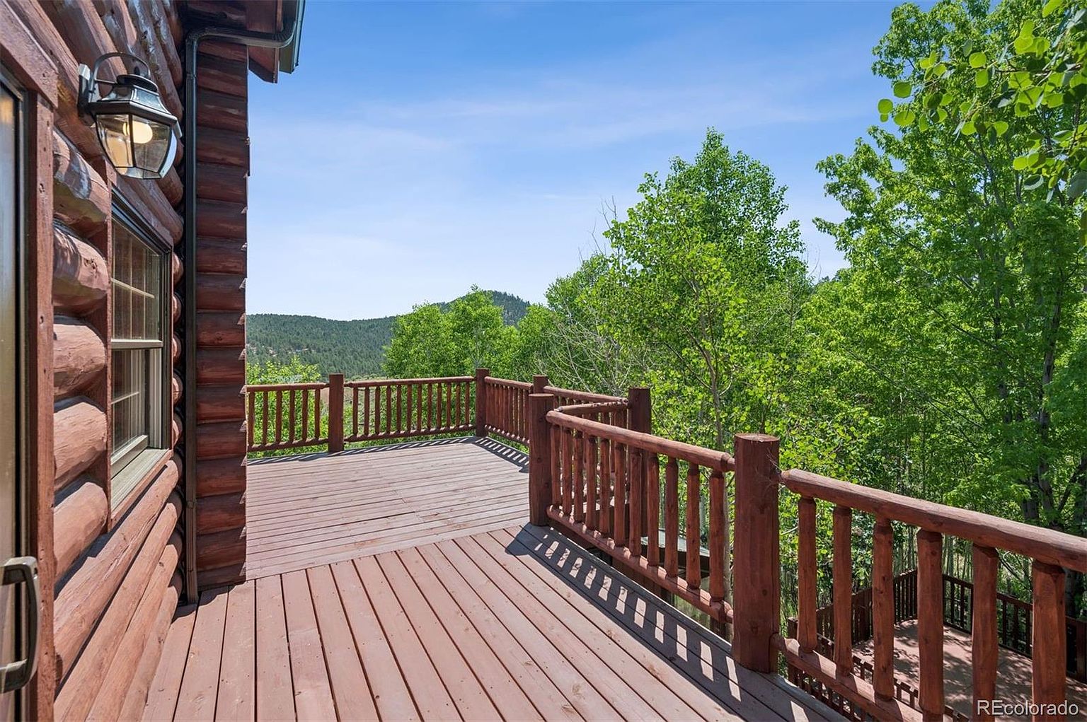 This image showcases a spacious wooden deck attached to a log cabin-style home. The deck features a sturdy wooden railing and offers a scenic view of the surrounding trees and distant hills. The warm tones of the wood and the natural setting create a rustic and inviting atmosphere, perfect for outdoor relaxation and entertaining.