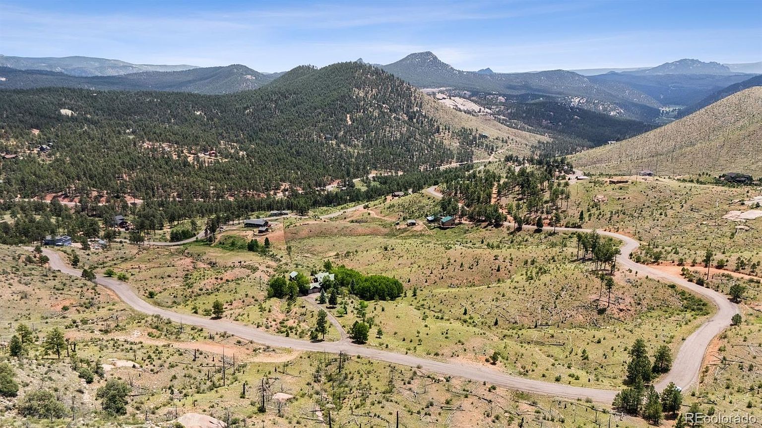 This aerial view showcases a sprawling property nestled in a mountainous landscape. A winding road leads to a cluster of homes surrounded by lush greenery and open fields. The scene evokes a sense of tranquility and seclusion, highlighting the property's natural setting and potential for privacy.